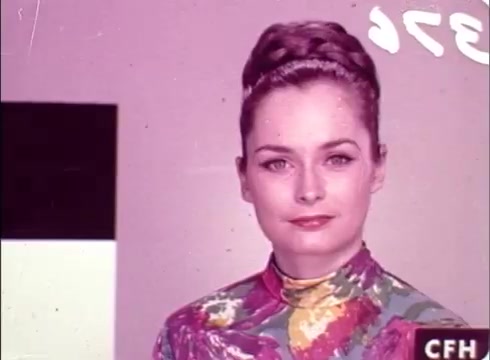 The still features a woman with a glossy hairstyle and colorful floral clothing, set against a muted background. She appears to be part of a screen test or promotional shoot, with a section of a color chart visible in the frame’s lower left-hand corner. The overall aesthetic reflects the vibrant style of the era. The image may show some signs of deterioration typical of 16mm prints.