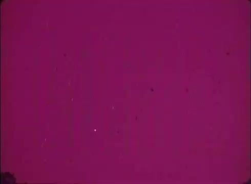 The still features a solid magenta background with visible grain and specks, indicative of a lab reject from a 16mm print. In the lower left-hand corner, there is a faint object, but the overall image is dominated by the deep coloration and texture of the film.