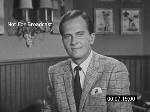 The still features Pat Boone, dressed in a light plaid suit with a tie, seated in a well-lit room. He appears earnest as he speaks directly to the camera, promoting the benefits of savings bonds during the Vietnam War era. The background includes wall decor and hints of a cozy setting, emphasizing the personal appeal of his message. He mentions President Johnson, aligning his appeal with the administration's financial initiatives. The footage is marked 'Not For Broadcast,' indicating it was likely created for promotional or educational purposes rather than public airing.