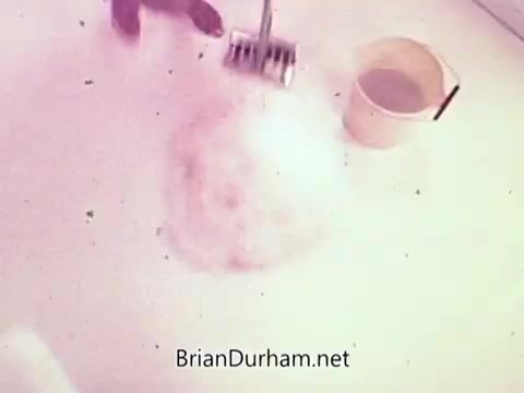 The still from the musical TV commercial for Tomorrow's Lestoil features a close-up of a cleaning scene. A hand is seen using a cleaning tool, likely a scrubber or brush, on a surface that is covered with foam and suds. Next to the cleaning action, there's a bucket, presumably containing Lestoil cleaner. The overall setting conveys a focus on effective cleaning, likely enhanced by the upbeat musical elements typical of commercials from that era.