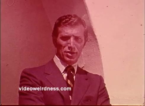 The still features Joe Campanella, dressed in a formal suit with a striped tie, standing confidently against a light-colored architectural backdrop. The image has a vintage aesthetic typical of 16mm film, characterized by warm tones and slight graininess, enhancing the nostalgic feel of the commercial for the Unity Church.