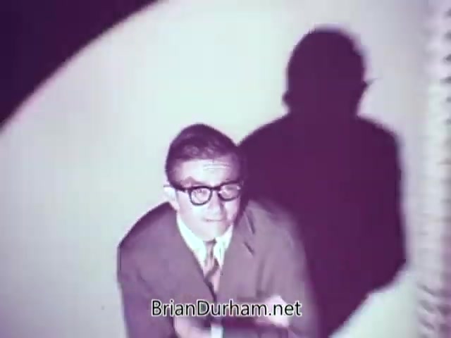 The still from the Heinz Baby Food commercial features a man with glasses, likely portraying a humorous or engaging character. He appears beaming toward the camera, with a bright background that suggests an upbeat atmosphere. His shadow is cast dramatically behind him, adding a playful element to the scene. The overall tone hints at a lighthearted promotion of the baby food product.