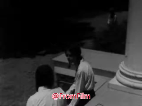 The still from the 1968 PSA showcases a scene at an orphanage, highlighting a sense of isolation. It features two children sitting on a porch, dressed in similar clothing, indicative of the era. The focus is on their expressions, which convey a mix of introspection and solitude. The atmosphere appears somber, reinforcing the message of the voice-over about the little girl's struggles with shyness and feeling alone among her peers. This visual aids in evoking empathy for the children's situation.
