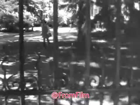 The still from 'PSA From Guideposts Magazine - 1968' depicts a young girl standing outside an orphanage. She appears isolated and alone, with a sense of shyness that sets her apart from others. The scene is framed through a wrought-iron gate, suggesting both physical and emotional barriers. The surrounding environment is lush, indicating the contrast between her loneliness and the vibrancy of the world around her. The overall tone evokes a poignant reflection on childhood and the challenges faced by those in orphanages.