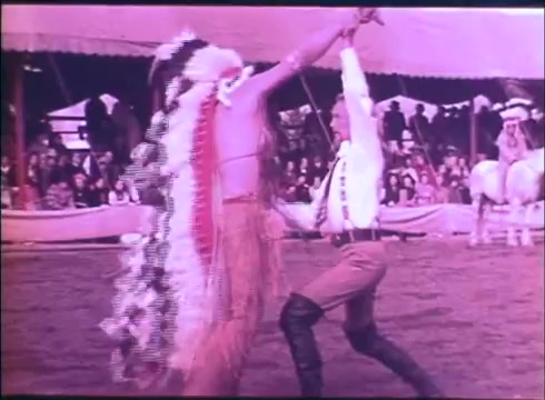 The still from the trailer of 'Buffalo Bill and the Indians' depicts a lively scene featuring two characters engaged in a dance. One performer is clad in traditional Native American attire, complete with a feathered headdress, while the other is dressed in a shirt and trousers, suggesting a contrasting cultural background. The setting appears to be an outdoor event or performance, with an audience visible in the background, all under a large tent. The colors have a vintage quality, characteristic of 16mm film.