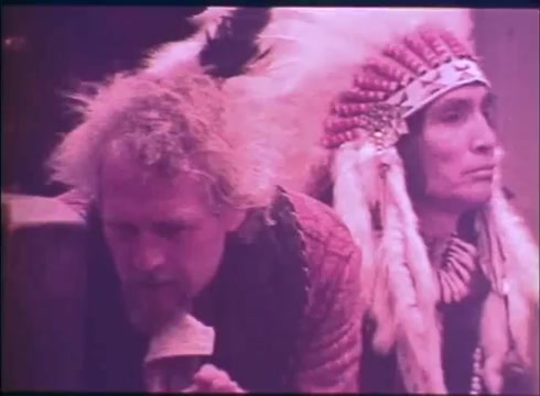 The still from the trailer of 'Buffalo Bill and the Indians' features two men: one with curly, light-colored hair dressed in period attire, and the other, an Indigenous man wearing traditional regalia and a feathered headdress. They appear to be engaged in a serious moment, highlighting themes of cultural interaction and conflict. The image has a pinkish hue, contributing to a distinct visual style.