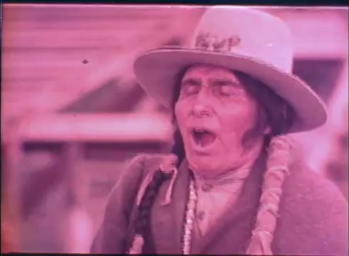 The still from 'Buffalo Bill and the Indians' trailer features a man wearing a large, wide-brimmed hat and traditional attire. He has long hair, a braid, and appears to be in mid-expression, likely vocalizing or singing. The colors in the image have a warm, muted tone, contributing to the historical ambiance of the scene. The background suggests a rustic setting that aligns with the themes of the film.
