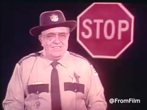 In this still from a 16mm print of a 1970s PSA addressing the energy crisis, a smiling, bespectacled sheriff stands prominently in front of a bright red stop sign that reads 'STOP.' Dressed in a classic sheriff's uniform, complete with a hat, he appears to convey a sense of authority and friendliness. The PSA likely aims to promote the use of fluorescent lighting as a means to save energy during a time of crisis. The visual style is characteristic of 1970s educational films, with vibrant colors and straightforward messaging.