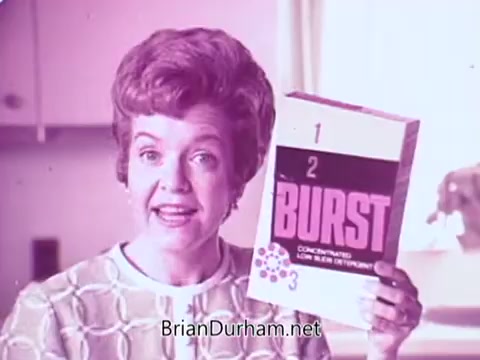 The still features a woman with a vintage hairstyle, smiling and holding a box of 'Colgate Burst' low suds laundry detergent. The packaging prominently displays the name 'BURST' alongside a few colorful dots, suggesting a vibrant and effective product. The background appears to be a bright kitchen, contributing to a cheerful and homey atmosphere. The overall tone is nostalgic and promotional, emphasizing the product's features.
