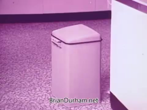 The still features a simple kitchen scene with a low, square trash can prominently displayed on a patterned floor. The overall color tone appears to be a soft pink, highlighting the product's branding. The surrounding environment suggests a mid-century aesthetic typical of the era when the advertisement was likely produced, aimed at showcasing the convenience of low suds laundry detergent.