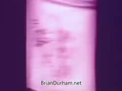 The still from 'Colgate Burst - low suds laundry detergent' shows a close-up, slightly blurred view of a detergent container. The color scheme leans towards a pinkish hue, highlighting the packaging and brand. The focus seems to be on the product's name and essential features, emphasizing its low suds formula. The image captures the essence of marketing from the era, showcasing detergent in a visually appealing manner.