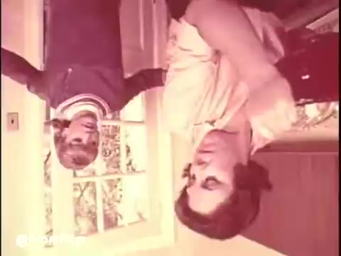 The still from 'Bounty Commercial Threaded Backwards...' features a scene in a domestic setting, likely depicting a mother and child interaction. The child, dressed casually in a jacket with a striped collar, appears to be engaging with the woman, who is dressed in a light, flowing top. The background suggests a cozy home environment, with natural light filtering in through a window. The film has a vintage aesthetic typical of 16mm prints, lending it a nostalgic feel. The overall mood may evoke warmth typical of family-oriented commercials from the era.