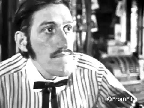 The still from the 1970s Jello commercial showcases a man with a prominent mustache and a striped shirt. The image is in black and white, resulting from a faded color treatment originally from a 16mm print. The man's expression appears contemplative or curious, focused on something off-camera. The background seems cluttered, typical of vintage commercial settings. The overall vibe evokes a nostalgic feeling, characteristic of advertisements from that era.