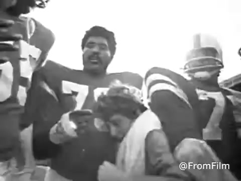 This still from a 1970s Jello commercial features a group of football players, with one prominent player in a jersey number 77. The scene captures a celebratory or casual moment, likely highlighting the fun and joy associated with Jello. The image has been converted to black and white, giving it a vintage look that reflects its origins from a 16mm film print. The faded colors suggest a nostalgic vibe, typical of commercials from that era.