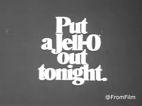 The still from the 1970s Jello commercial features bold, playful text that reads, 'Put a JELLO out tonight.' The design captures the vintage style of the era, with a font that is both inviting and fun. The overall look is in black and white due to the faded color treatment from the 16mm print, giving it a nostalgic feel. It's a simple yet effective call to action, typical of commercials from that time period.