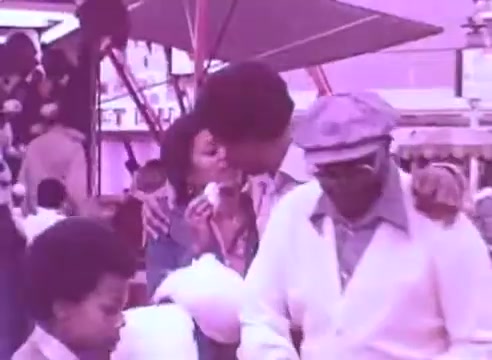 The still features a group of people in a vibrant outdoor setting, likely at a fair or festival. A couple shares a tender moment, while an older man dressed in a light-colored suit and hat engages with a young boy nearby. The scene conveys a sense of community and familial bonds, underscored by the health-related message of taking blood pressure medication. The color tone adds a nostalgic feel, enhancing the warmth of the moment.