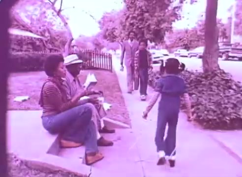 The still from 'Do it for them' - take your blood pressure medication 3 shows a street scene with a few individuals. A woman and a man are sitting on the sidewalk, engaged in conversation and sharing food. Nearby, a child walks past them, appearing playful and carefree. In the background, additional figures are walking along the sidewalk, and parked cars line the street. The overall atmosphere is community-oriented, highlighting intergenerational interactions. The color tone gives the image a nostalgic feel.