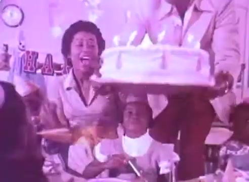 The still features a cheerful scene, likely a birthday celebration. A woman, joyfully smiling, holds a decorated cake adorned with candles, while a child sits in front, also excited. Other children are present, suggesting a festive atmosphere. The colorful decorations in the background enhance the celebratory mood, emphasizing the importance of family and moments worth cherishing, aligning with the theme of taking blood pressure medication for loved ones.