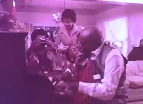 The still from 'Do it for them - take your blood pressure medication 3' depicts a warm, family-oriented scene at home. An elderly man is playing a piano while surrounded by family members, including a smiling child and adults engaging joyfully. The atmosphere is lively and affectionate, emphasizing the importance of family connection in health discussions. The overall color tone has a vintage feel, likely due to the 16mm film quality.