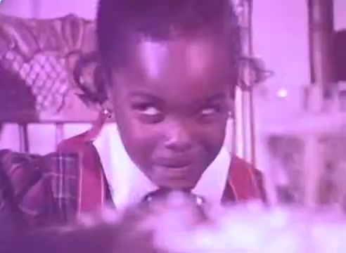 The still from 'Do it for them – take your blood pressure medication 3' features a young child with an expressive face. The child appears to be looking mischievously or playfully, suggesting a moment of engagement or curiosity. The colors are warm and somewhat muted, enhancing the vintage feel of the 16mm format. The background indicates a domestic setting, likely during a family gathering. Overall, it conveys a sense of innocence and the importance of relationships in health messaging.