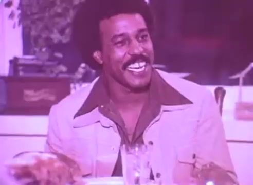 The still from 'Do it for them - take your blood pressure medication 3' features a smiling man with an afro and a stylish outfit, seated at a dining table. The background suggests a warm, inviting atmosphere, possibly during a family gathering or meal. The color tone has a vintage feel, likely from the time the film was made. The scene conveys positivity and connection, emphasizing the message of caring for one's health for the sake of loved ones.