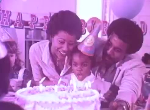 The still depicts a joyful birthday celebration, featuring a group of people, including a woman and a man, who are smiling and leaning toward a young child in a party hat. The child, also wearing a party hat, is seated in front of a decorated cake with lit candles. Colorful decorations and a 'Happy Birthday' banner can be seen in the background, enhancing the festive atmosphere. The scene conveys warmth, family interaction, and the importance of celebrating moments together, likely reinforcing the message behind taking blood pressure medication for loved ones.