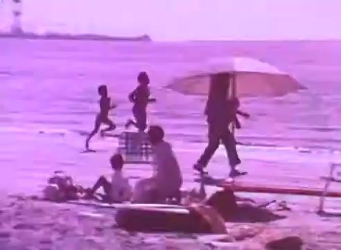 The still depicts a beach scene with a soft purple hue. In the foreground, a few people are relaxing on the sand, while a child is seated. To the side, a figure is walking with an umbrella, and in the background, several individuals can be seen running along the water's edge. The overall mood is calm and leisurely, emphasizing a sense of community and the importance of taking care of one’s health in a vibrant setting.