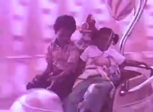 The still depicts two children and a clown on a ride, likely at a fair or amusement park. The setting has a playful, whimsical feel, enhanced by the bright colors and cheerful expressions, emphasizing enjoyment and innocence. The context of medication suggests a focus on health and well-being, advocating for taking blood pressure medication for the sake of loved ones.