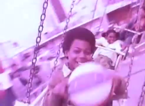 The still features a young boy with an afro, joyfully playing with a beach ball while on a swing ride. The background is blurred, suggesting motion and excitement, indicative of a funfair or amusement park setting. The overall color tone is a vivid pinkish hue, contributing to a nostalgic and cheerful atmosphere. The context suggests a focus on maintaining health and well-being, encouraging viewers to take their blood pressure medication for the sake of family and loved ones.