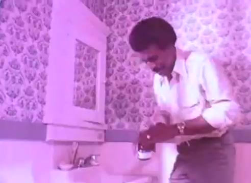 The still from 'Do it for them - take your blood pressure medication 3' depicts a man in a bathroom, dressed in a white shirt and light trousers. He is focused on opening a bottle of medication. The surroundings feature pastel-colored wallpaper and a mirror, emphasizing a personal moment in the context of health and self-care. The overall atmosphere conveys a sense of routine and responsibility regarding blood pressure management.