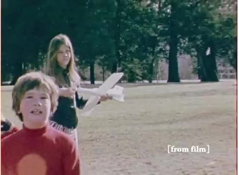 The still is from a 16mm film, dating back to 1972, featuring a toy commercial for the 'Flying Submarine' by Goodtone. In the scene, a young boy in a red sweater appears animated, while a girl, possibly his sibling or friend, is holding what seems to be a model airplane. The backdrop consists of a park with trees, suggesting an outdoor setting ideal for showcasing the toy. The film retains a nostalgic quality, captured in a vibrant color palette characteristic of that era. The soundtrack accompanying the footage is noted as the 'good' version, contrasting with another marked 'DO NOT USE.'