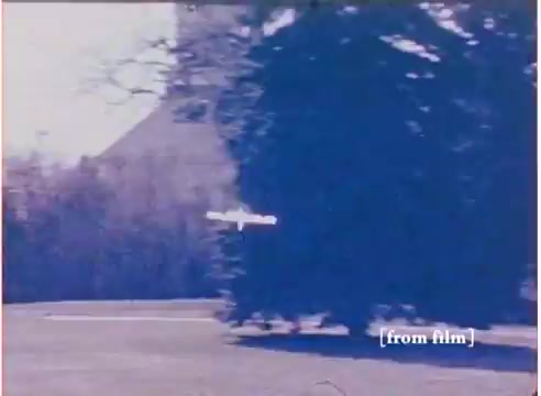 The still from the 16mm toy commercial features a surreal scene showcasing the 'Flying Submarine' toy. The image captures a bright, sunny day with a blurred background of trees and a structure, suggesting an outdoor setting. The toy appears to be gliding or hovering in mid-air, emphasizing its unique flying capability. The film has a vintage quality, characteristic of 1972, and the still includes the notation “[from film]” at the bottom, indicating its source. The separate soundtrack, labeled as the 'good' version, adds to the commercial's authenticity.