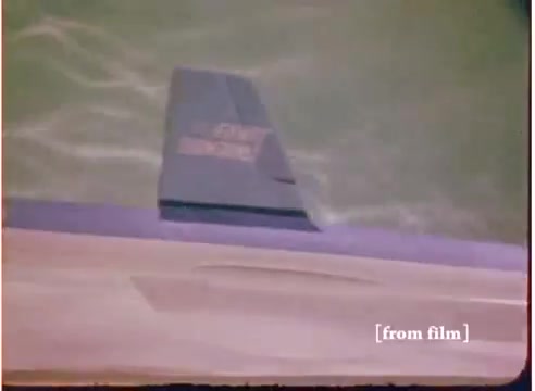 The still from the 16mm toy commercial depicts a close-up of a 'flying submarine' toy. The image captures a sleek, gray model, partially submerged in water, showcasing its streamlined design. The surrounding water appears rippled, suggesting that the toy is in motion. The film has a vibrant, slightly faded color palette typical of the early 1970s. The accompanying soundtrack is noted as the 'good' version, indicating a specific emphasis on quality for this presentation, contrasting with another version labeled 'DO NOT USE.'