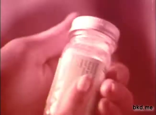 The still features a close-up of a hand holding a glass jar of Mentholatum, with the label partially visible. The lighting gives it a warm, nostalgic feel typical of 1970s commercials. The focus on the jar emphasizes the product, suggesting its use for soothing cold symptoms. The image captures a sense of comfort and care associated with the brand.
