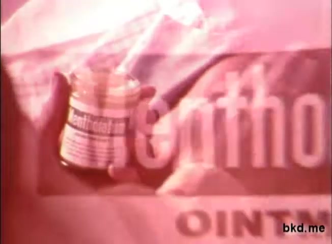 The 16mm still from the 1970s Mentholatum for colds commercial features a close-up of a person's hand holding a small jar of Mentholatum ointment. The packaging is prominently displayed, showcasing the brand name. The background has a soft focus, creating a warm, nostalgic atmosphere typical of the era's advertising. The overall color tone leans towards muted pastels, conveying a sense of comfort associated with cold relief.