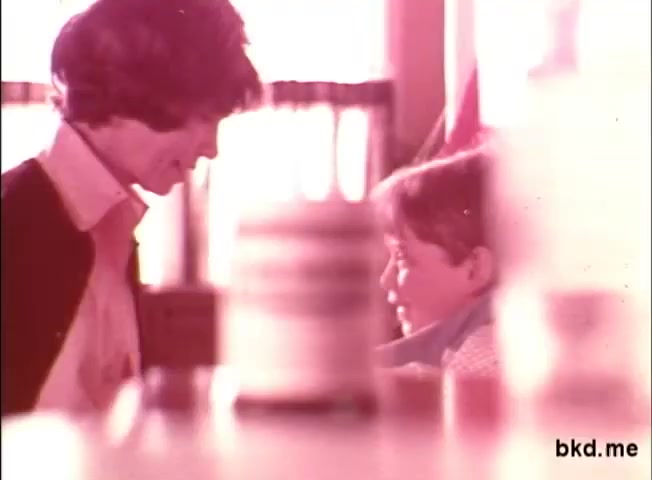 The still captures a warm, intimate moment between a woman and a young boy, likely indicative of a mother-child relationship. The woman appears to be gently engaging with the boy, who looks up at her. The soft focus and pastel color tones evoke a nostalgic atmosphere typical of 1970s commercials. In the background, there are various items, possibly medication, hinting at the theme of treating colds with Mentholatum.