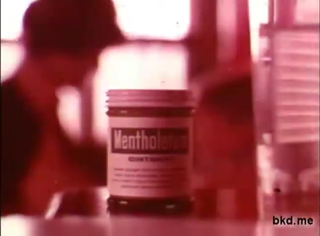 The still features a jar of Mentholatum prominently displayed in the foreground, showcasing its label. The background is slightly blurred, suggesting a dynamic environment, possibly indicating a home or pharmacy setting. The warm, faded color palette typical of 1970s film adds a nostalgic ambiance, emphasizing the product’s vintage appeal.