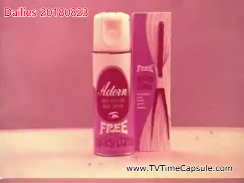 This 16mm still from 'dailies 20180823-scott-towels-etc' features a spray can of Adorn, prominently displaying the word 'FREE' on both the can and the accompanying box. The overall color palette is soft pink, suggesting a vintage or retro aesthetic. The product appears to be part of a promotional display aimed at showcasing its features.
