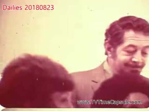 The still appears to be a vintage shot from a film or video, possibly a behind-the-scenes or candid moment. It features a man in a suit smiling alongside people with curly hair, captured in a warm, nostalgic tone typical of 16mm film. The context suggests it might relate to a personal or informal gathering.