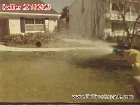 The still features a person in a yellow shirt spraying water in a yard, using a garden hose. The background includes a residential house and a lawn, suggesting a casual outdoor setting, likely during a warm day. The image conveys a sense of playfulness and everyday life.