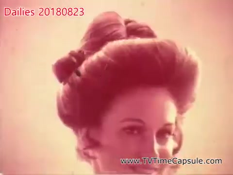 The still features a woman with a styled hairdo, prominently showcasing volume and elegance. The background is minimal, creating a focus on her expression and hairstyle. The color tone has a warm, nostalgic effect, reminiscent of vintage film aesthetics. This image likely captures a moment from a promotional shoot or a fashion-related dailies production.