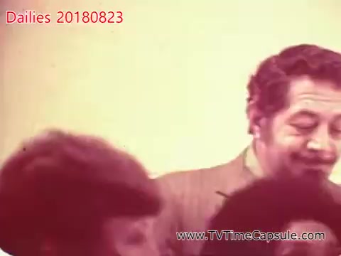 The still likely depicts a group of people engaged in a social interaction. A man is prominently featured, with a thoughtful expression, while others, possibly women, are partially visible in the foreground. The overall atmosphere appears informal, suggesting a candid moment captured during dailies. The image has a vintage aesthetic typical of 16mm film.