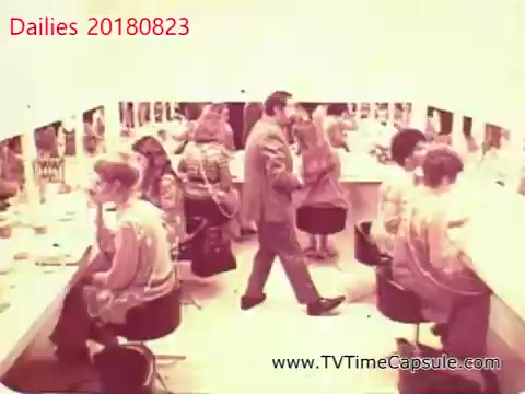 The still features a bustling makeup room, with multiple people seated at vanities applying makeup or preparing for a performance. A man in formal attire walks through the space, interacting with one of the women. The atmosphere suggests a behind-the-scenes look at a production, capturing a moment of preparation and camaraderie. The color tone indicates a vintage style, typical of 16mm film.