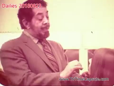 The still features a man in a suit interacting with a young girl, likely in a setting related to grooming or hairstyling. The scene conveys a sense of care or instruction, with the man's expression focused and engaging. The color and quality suggest it is a vintage 16mm film still.