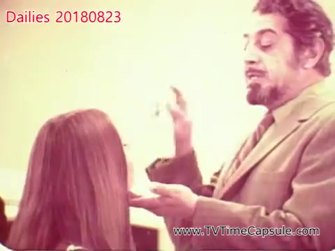 The still features a man in a suit interacting with a woman, who has long hair, in what appears to be a makeup or beauty setting. The man seems to be applying a product or demonstrating a technique, conveying an instructional or guiding role. The background is blurred, focusing attention on their interaction. The overall atmosphere suggests a casual yet professional environment, possibly from a film or promotional shoot.
