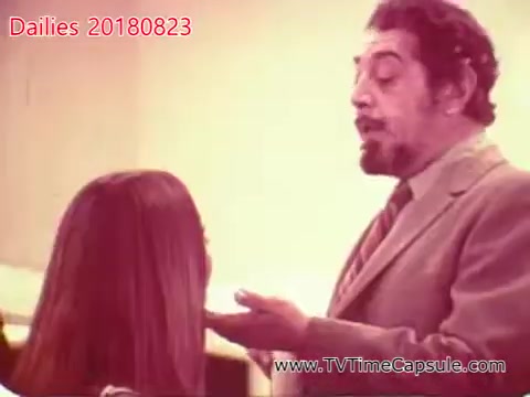 The still from 'dailies 20180823-scott-towels-etc' depicts a scene featuring a man in a suit engaging in a conversation with a woman. The man appears animated, possibly making a point, while the woman listens attentively. The setting has a soft-focus background, suggesting an indoor environment. Overall, the interaction seems to be focused and dynamic, hinting at a narrative development between the two characters.