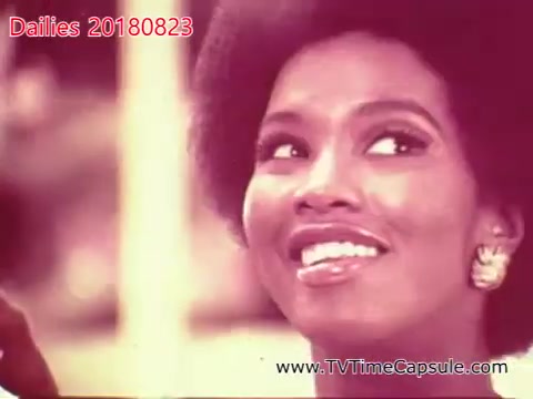 The still features a woman with an expressive smile, showcasing her natural hairstyle and elegant earrings. The background appears blurred, suggesting a candid moment. The overall tone is bright and inviting, capturing a joyful expression in a seemingly casual setting.