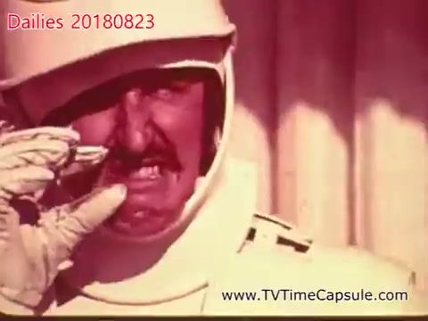 The still shows a man dressed in a white, futuristic outfit, possibly a space suit or costume, wearing white gloves. He has a mustache and appears to be making an exaggerated facial expression, suggesting frustration or an intense reaction. The background appears blurred, emphasizing the character's face and the dramatic moment. The color tone is warm, giving a nostalgic feel typical of 16mm film.