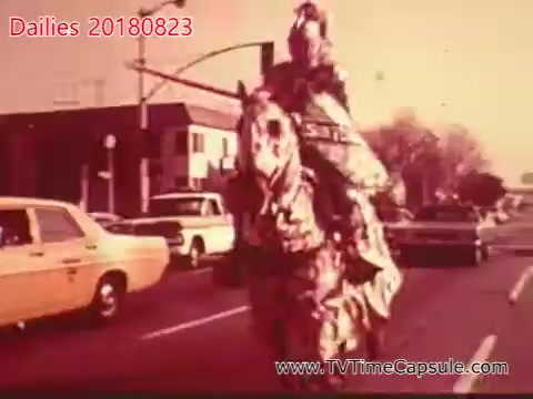 The still depicts a horse rider dressed in a distinctive costume, possibly historical or ceremonial, amidst a busy street filled with cars from the 1970s or 1980s. The background features buildings and traffic signals, indicating a suburban or urban setting. The overall color tone is warm and slightly faded, characteristic of 16mm film.