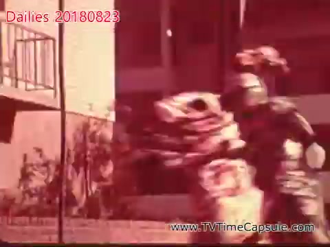 The still features a scene with performers in elaborate costumes, likely mimicking a traditional dance or cultural performance. The background shows a building and some greenery, indicating an outdoor setting. The overall tone is vibrant, suggesting a lively atmosphere.